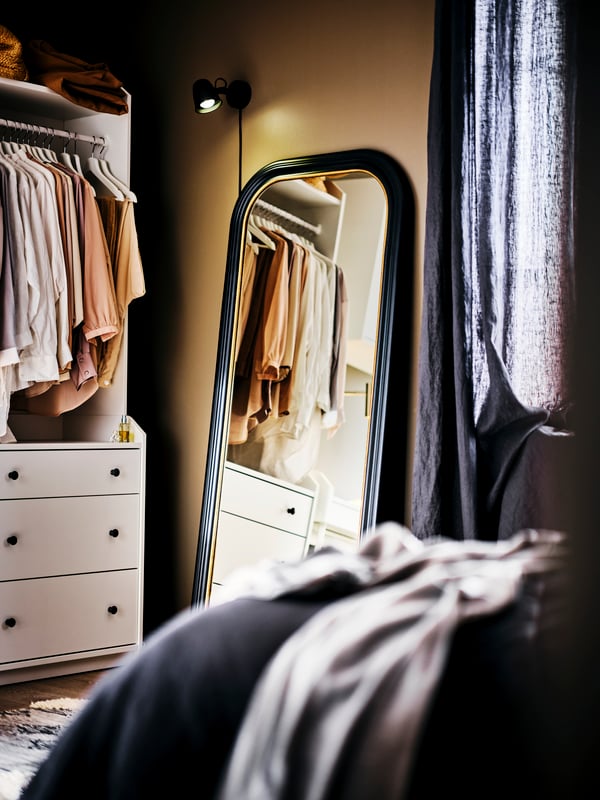 Ein ALMARÖD Spiegel lehnt an einer Wand mit einem offenen HAUGA Kleiderschrank daneben. Die Kleiderstange ist mit Kleidung gefüllt.