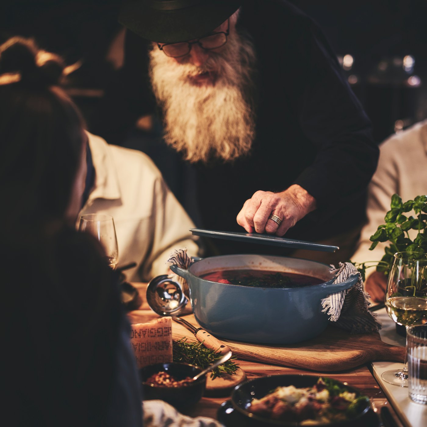 An elderly man with a long beard lifts the lid on a casserole dish while dinner guests seated at the table look on.