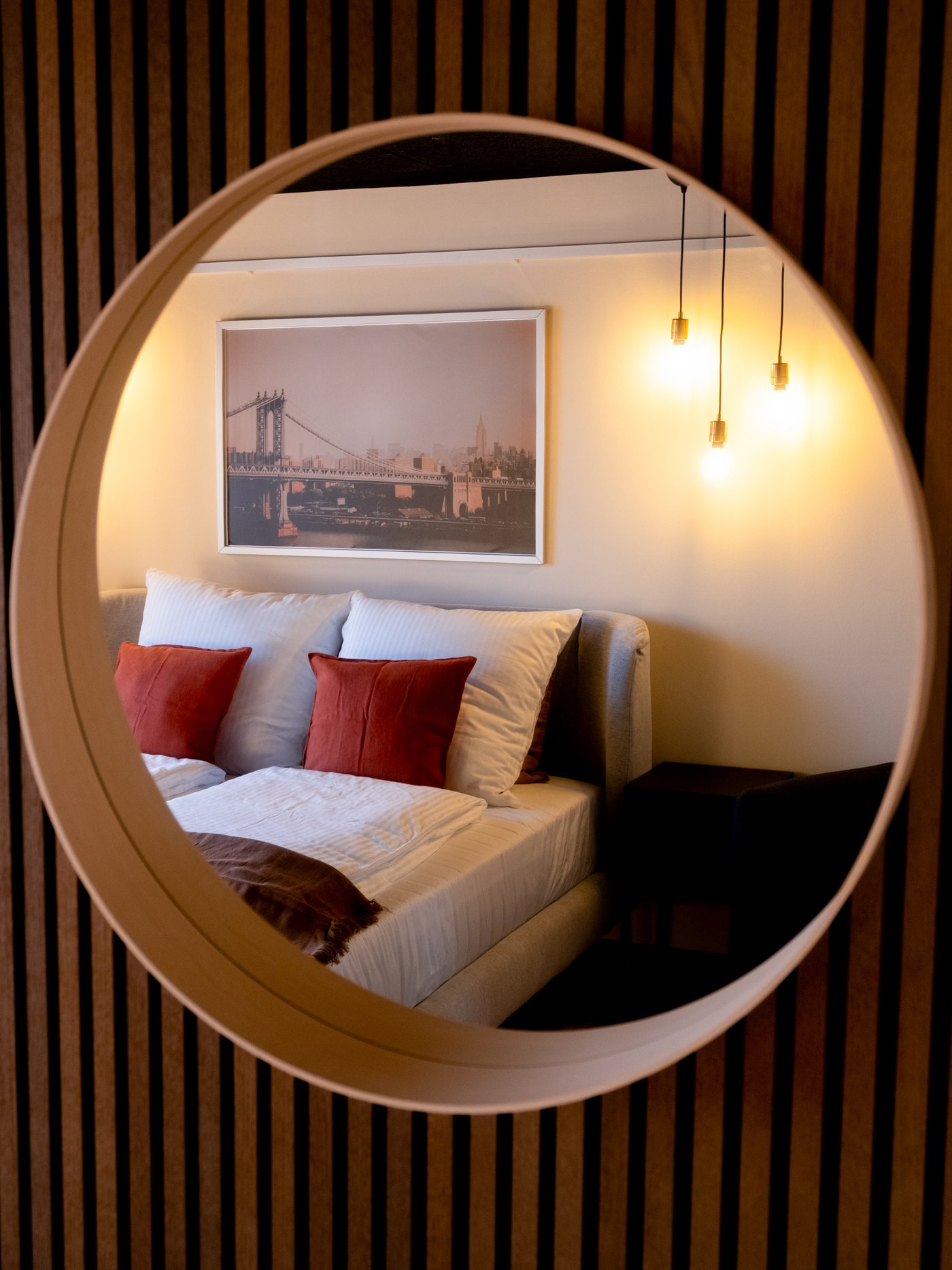 Bedroom reflected in a round mirror with bed, white pillows, rust-colored decorative pillows and warm lighting from hanging lamps.