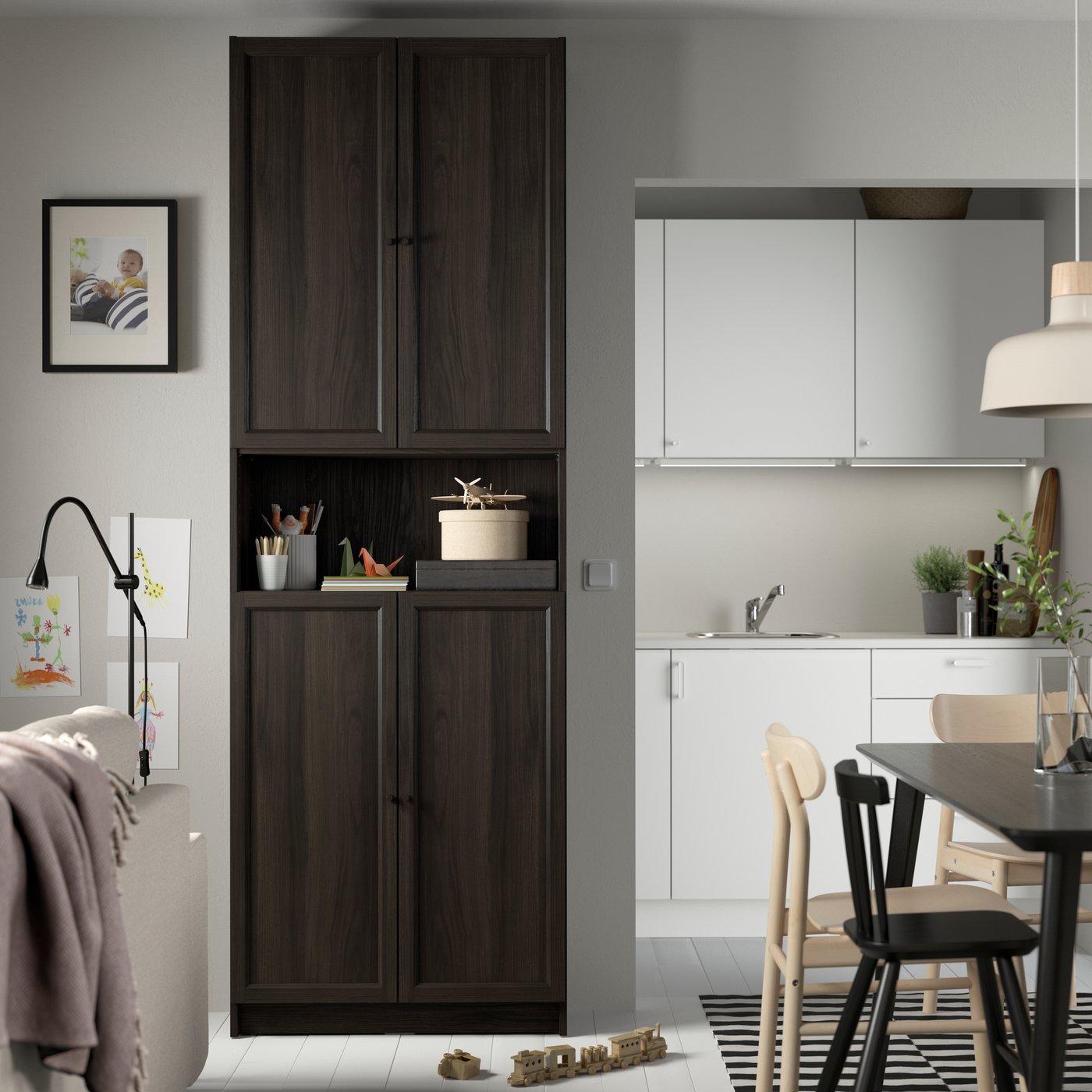A BILLY/OXBERG bookcase with doors in dark brown oak effect against a wall near a kitchen and dining area.