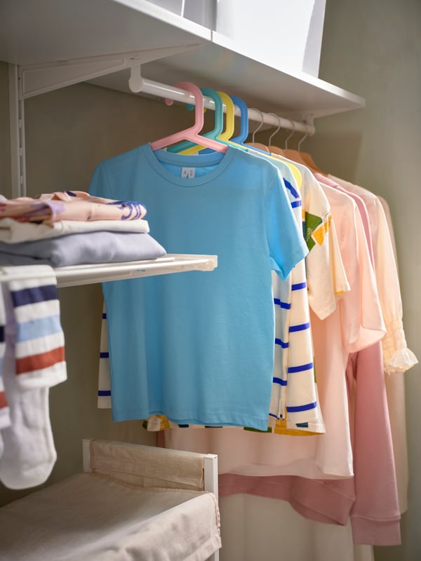 A storage room with a BOAXEL shelving unit featuring mixed-colour BAGIS children's clothes hangers holding children's clothing.