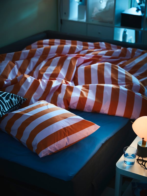 Una stanza in penombra con un divano letto ÄLVDALEN preparato con biancheria da letto MAJSMOTT. Una lampada NÖDMAST si trova su un vicino tavolino TINGBY.