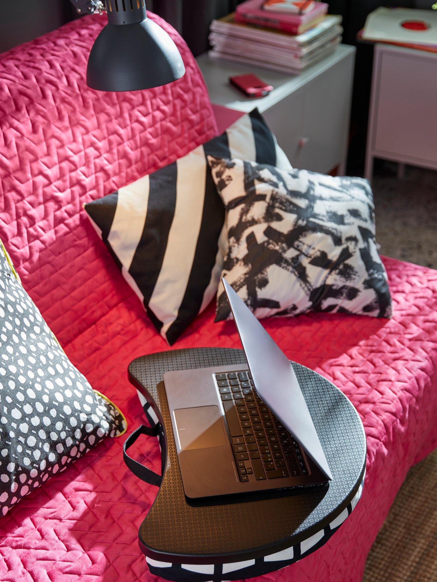 Una laptop negra sobre un soporte BYLLAN, apoyado en un sillón con forro rojo y cojines TURILL con diseños en blanco y negro.