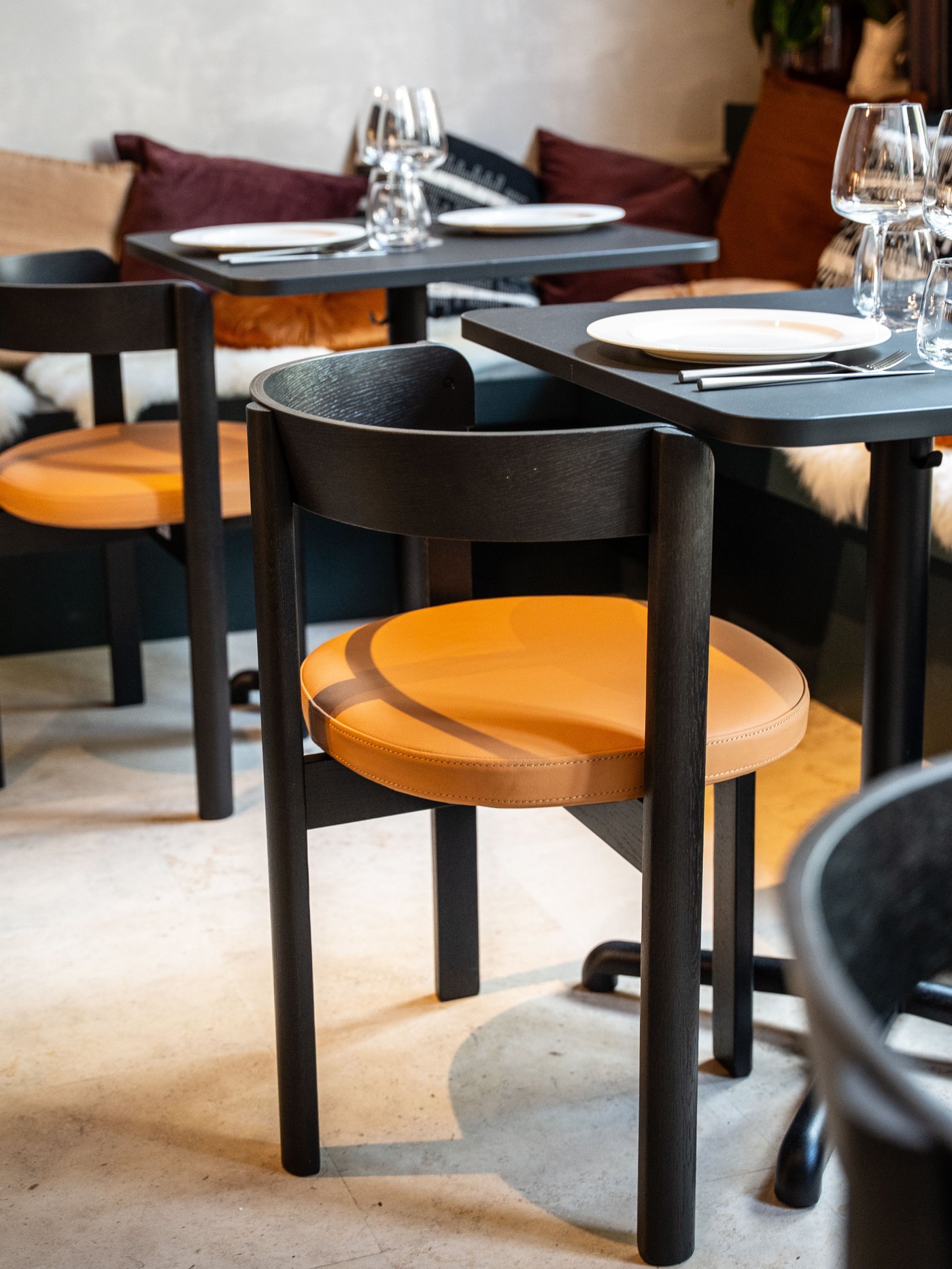 Zwarte tafels met oranje stoelen, gedekt met glazen en borden. Achteraan kussens op bank in warm, modern interieur met lichte vloer.