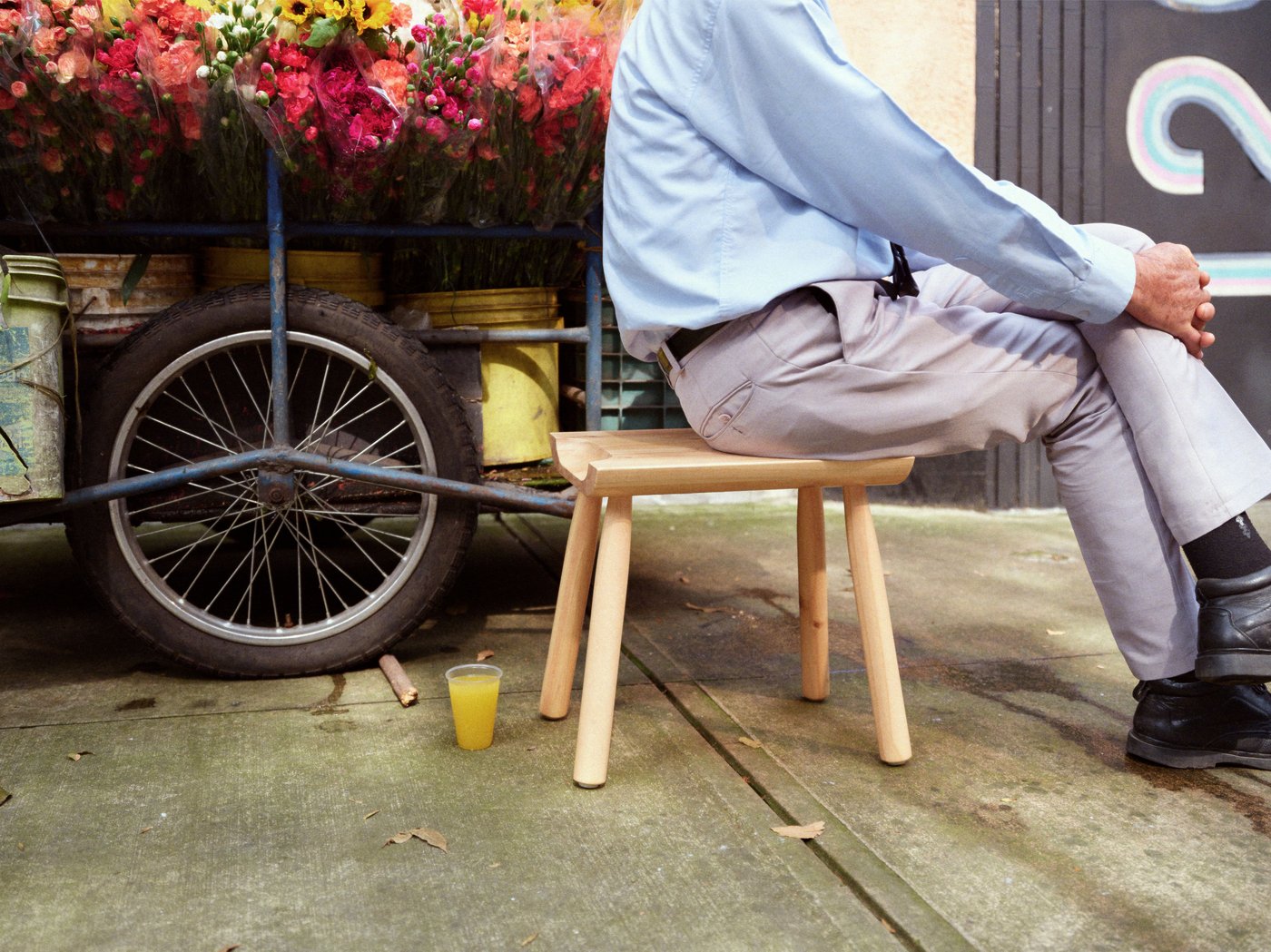 In uno spazio all'aperto, una persona è seduta a gambe incrociate su uno sgabello in legno. Sullo sfondo c'è una carrozza con dei fiori.