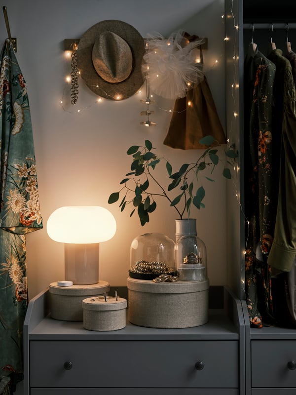 Commode grise décorée d'un éclairage chaleureux : lampe de table DEJSA allumée, guirlande lumineuse et objets décoratifs. 