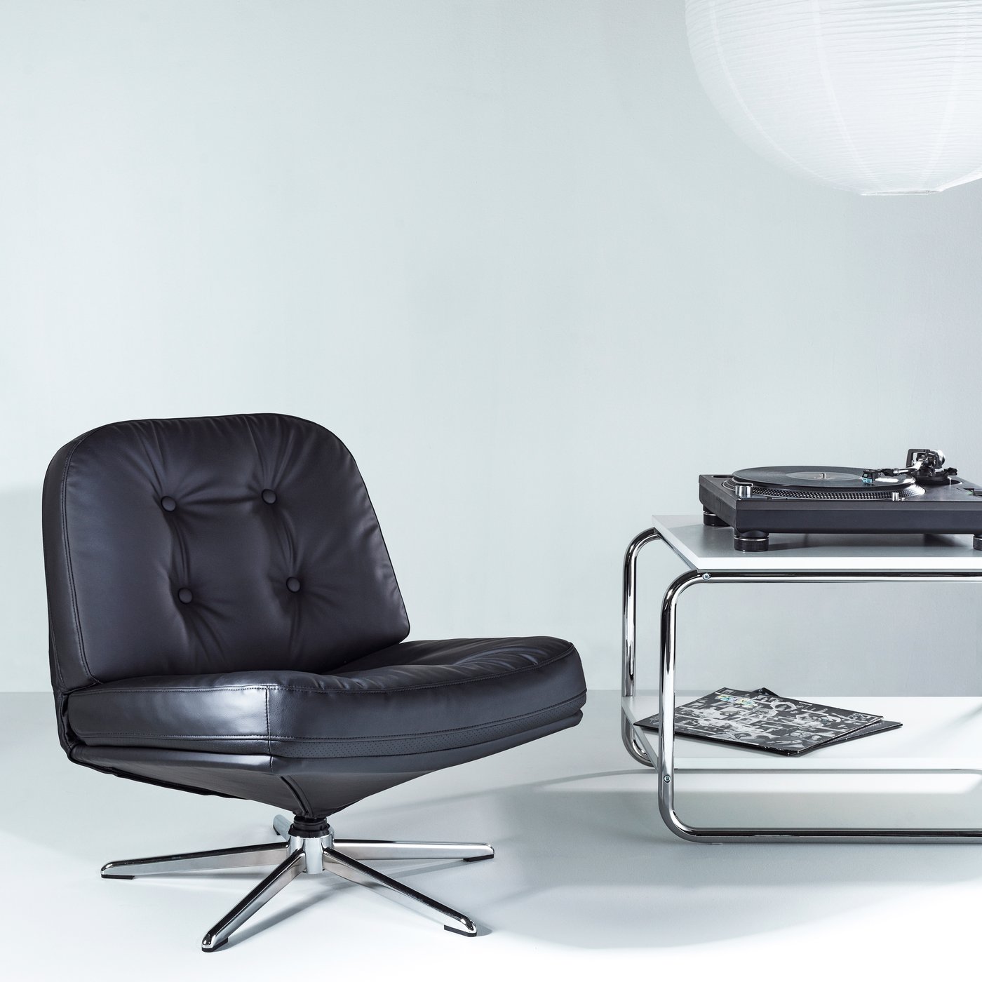 Black leather lounge chair with footrest beside an elegant chrome side table and turntable in a bright living room.