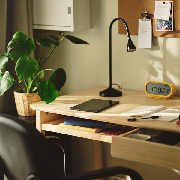 A workspace featuring a light bamboo BETESHÄST desk with a laptop and a clock on it, beside a green plant.