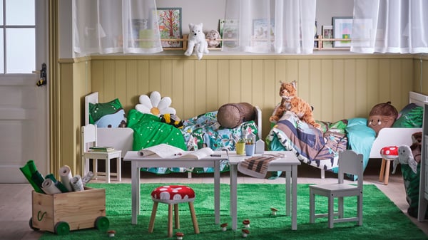 Es ist ein Kinderzimmer mit zwei Einzelbetten mit SKOGSDUVA Bettwäsche und SKOGSDUVA Stoffspielzeugen zu sehen. Auf zwei Kindertischen liegen Malutensilien.