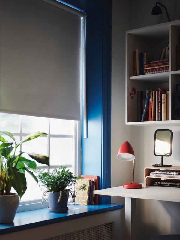 Lampe de travail à DEL ISNÅLEN rouge et couleur laiton bien en vue sur un bureau à côté d’une fenêtre habillée d’un store à enrouleur opaque FRIDANS blanc.