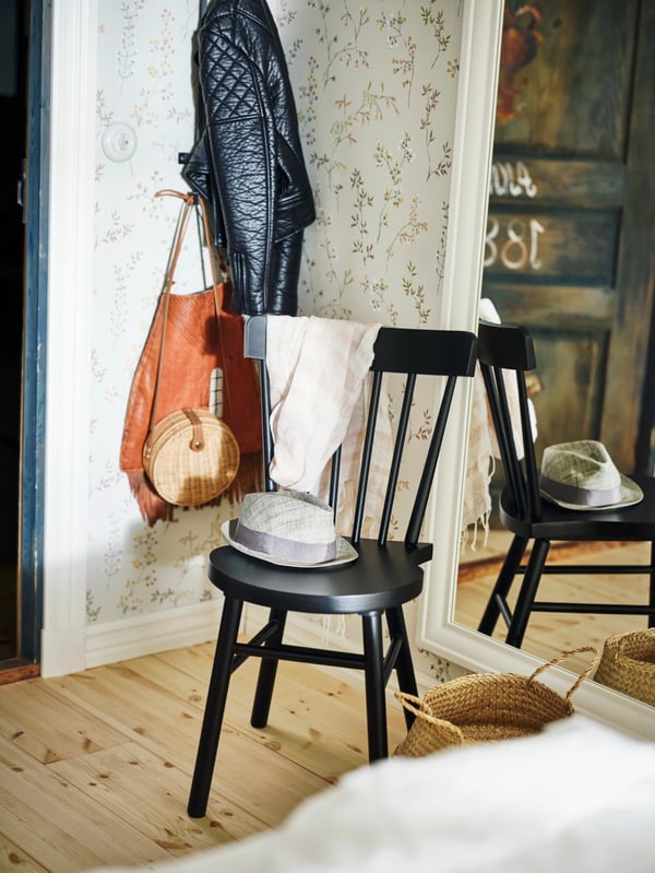 A black NORRARYD chair in front of a mirror and beside a basket on the floor, with a hat on the seat and a scarf on the back.