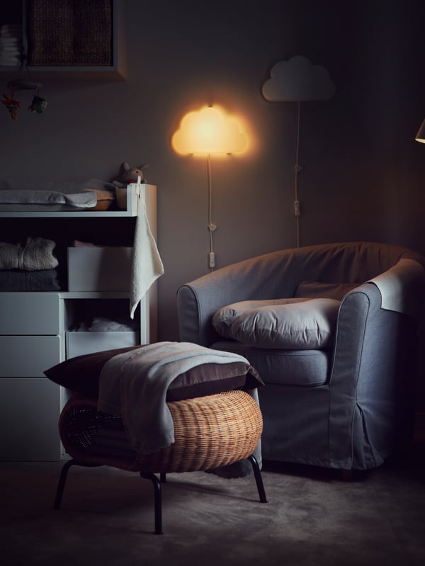 A cushions/blanket placed on a rattan GAMLEHULT footstool with storage in front of an armchair in a room lit by a night wall lamp.