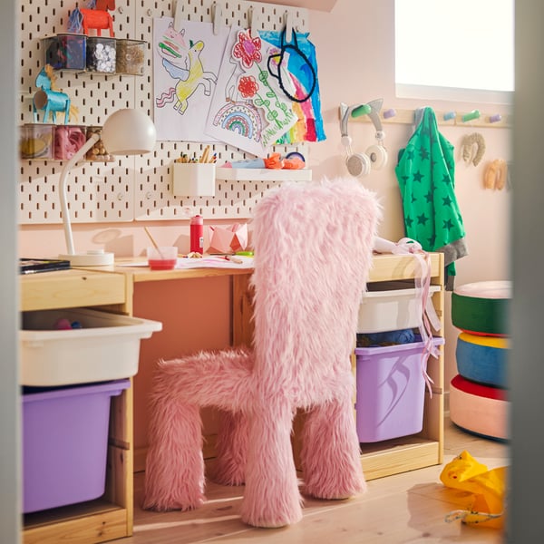 A children’s room with a pink faux fur GREJSIMOJS cover for children’s chair, a desk and storage units beside a pegboard wall.