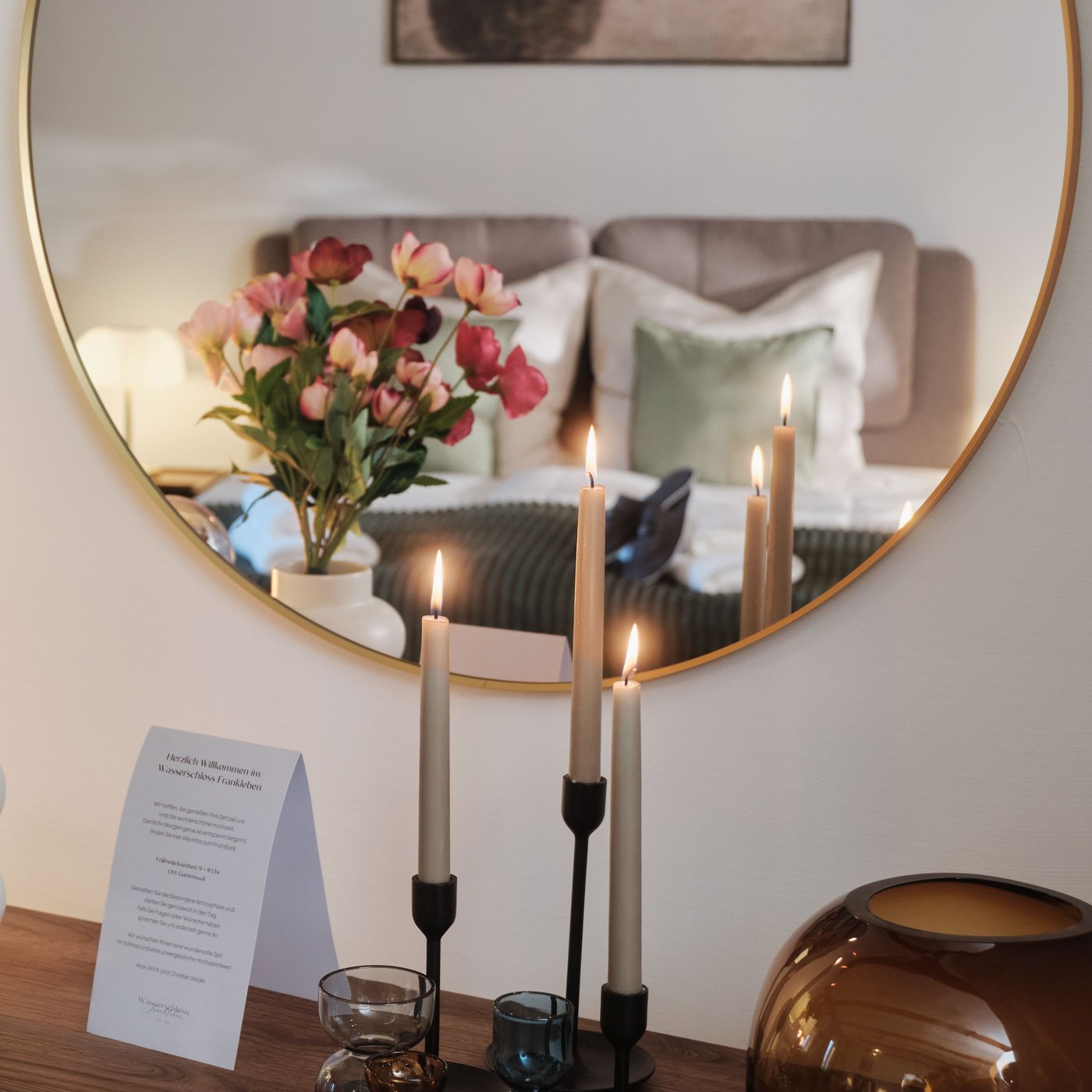 Byrå med tända ljus och blomsterbukett framför en rund spegel som reflekterar en bäddad säng i ett harmoniskt sovrum.