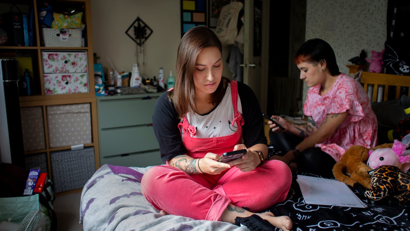 Two women sit on a bed, looking at their phones, surrounded by soft toys.