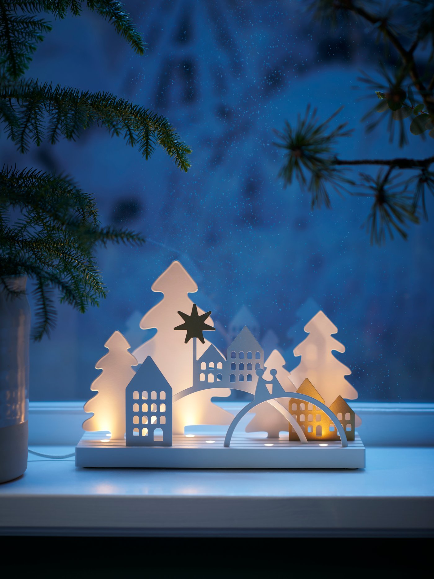 A STRÅLA decorative LED lamp that depicts a village scene is set on a windowsill on a frosty winter evening.