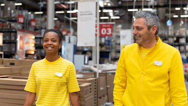 Two IKEA co-workers wearing a yellow IKEA unifome, walking through an IKEA store warehouse.