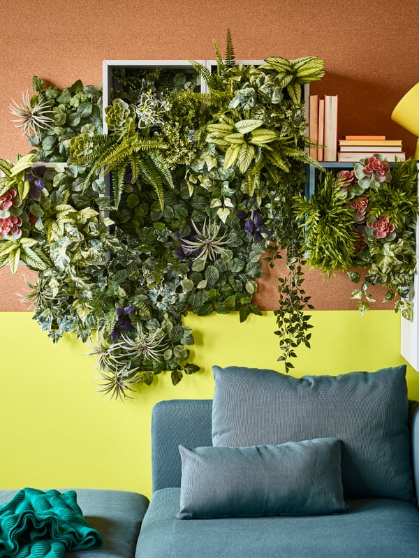 A living room with FEJKA artificial potted plant fern in plastic, above a sofa on a wall with fake plants.