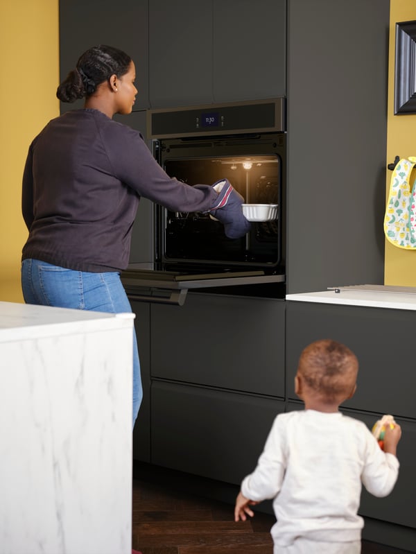A toddler looks on as a woman takes an oven dish out of a HJÄLTEBY wall oven in a kitchen with matt anthracite fronts.