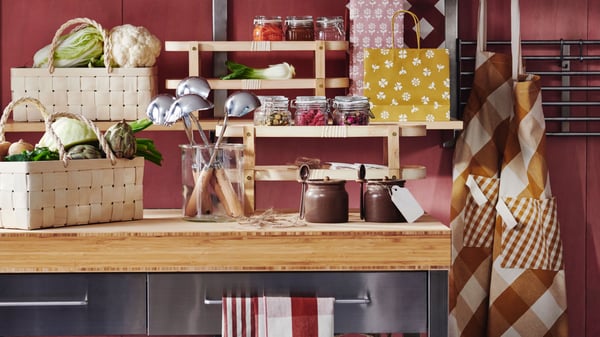 A kitchen with a RIMFORSA work bench made of stainless steel and bamboo, featuring various KÖSSEBÄR items such as jars, baskets, aprons, tea towels, and more.
