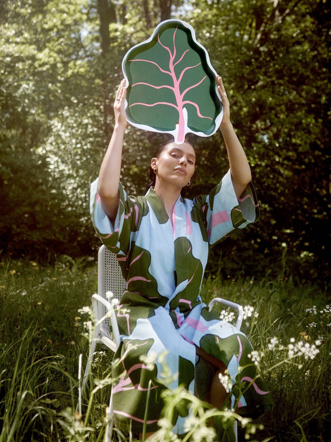 Seorang wanita yang memakai kimono BASTUA dengan corak daun sedang duduk di atas kerusi di padang rumput sambil mengangkat dulang berbentuk daun BASTUA.