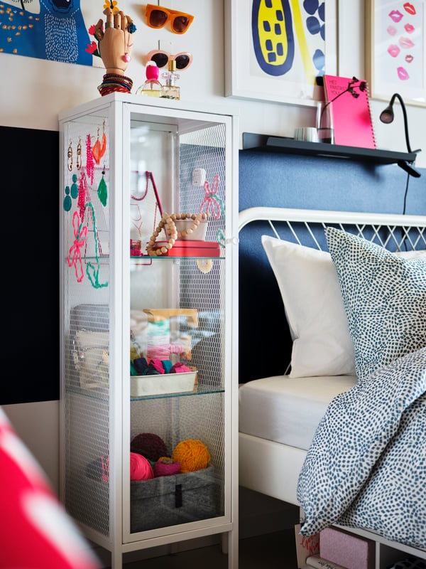 A white BAGGEBO glass door cabinet with items on display, beside a white NESTTUN bed with white/blue TRÄDKRASSULA bedlinen.