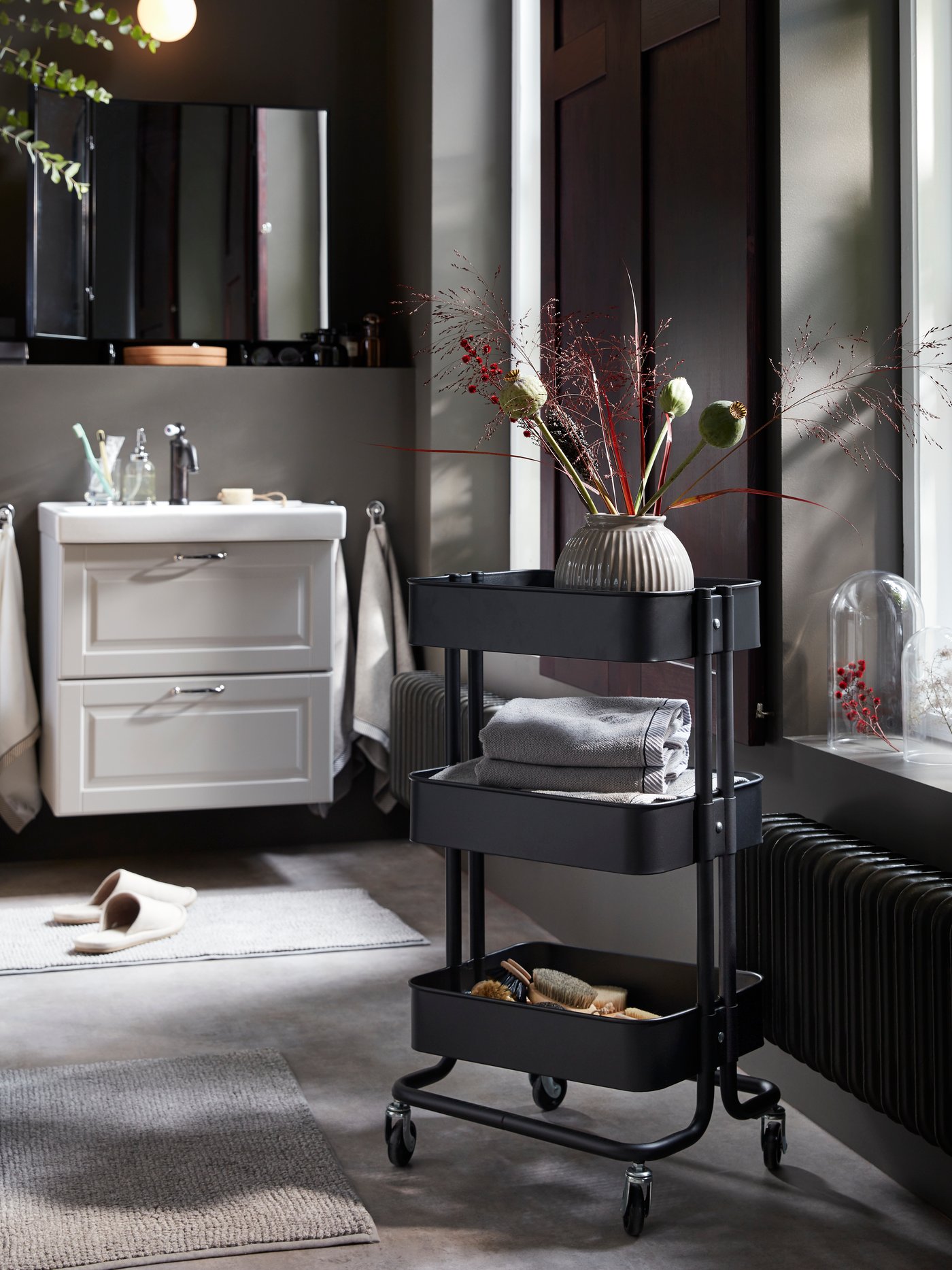 A black RÅSKOG trolley on castors with decorative flowers, hair accessories and folded grey towels on the shelves.