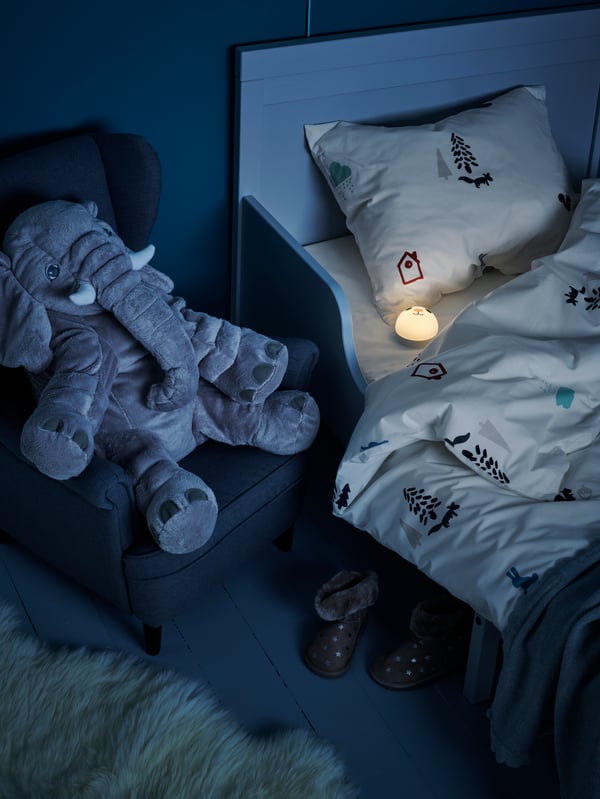 A BLÅVINGAD soft toy blue whale lies on a duvet with a BLÅVINGAD duvet cover on a KURA reversible bed.