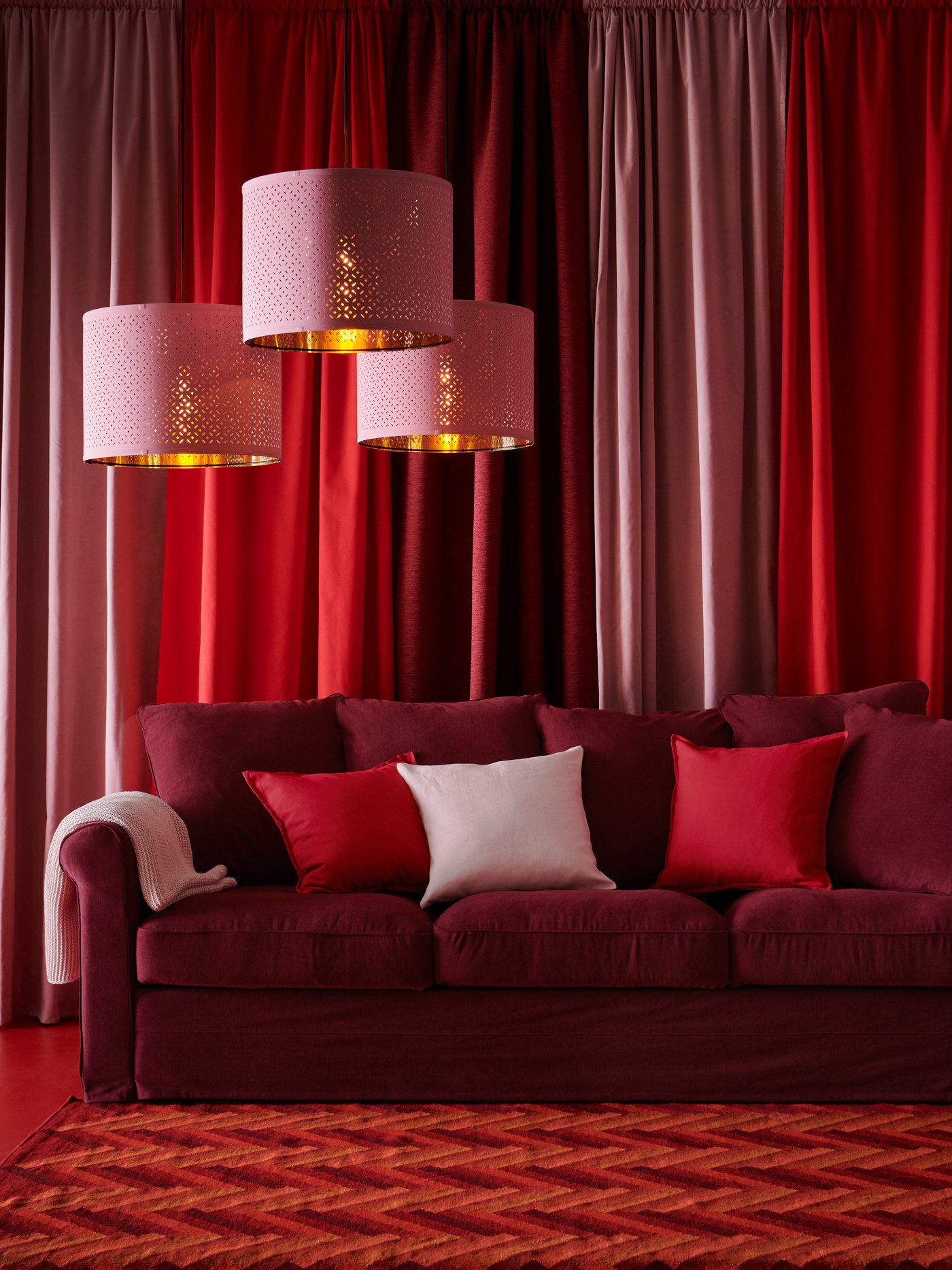 A collection of red pillows sit on a red couch on a wall carpet against red curtains on the wall. Three pendant lights hang to the left of the couch.