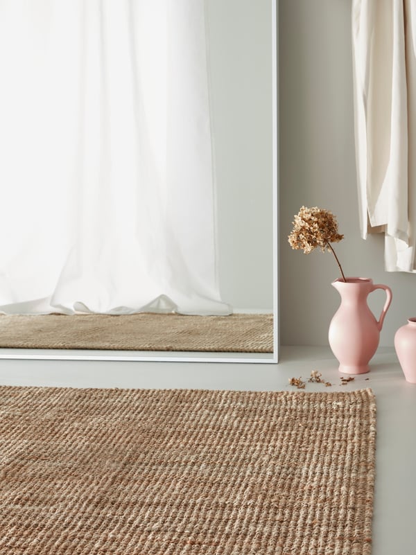 Un miroir HOVET repose contre un mur, à côté d’un tapis LOHALS en jute et de deux vases roses.