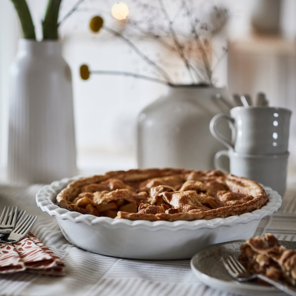 An off-white HÖSTAGILLE pie dish filled with apple pie sits on a stripey tablecloth, surrounded by cutlery and vases.