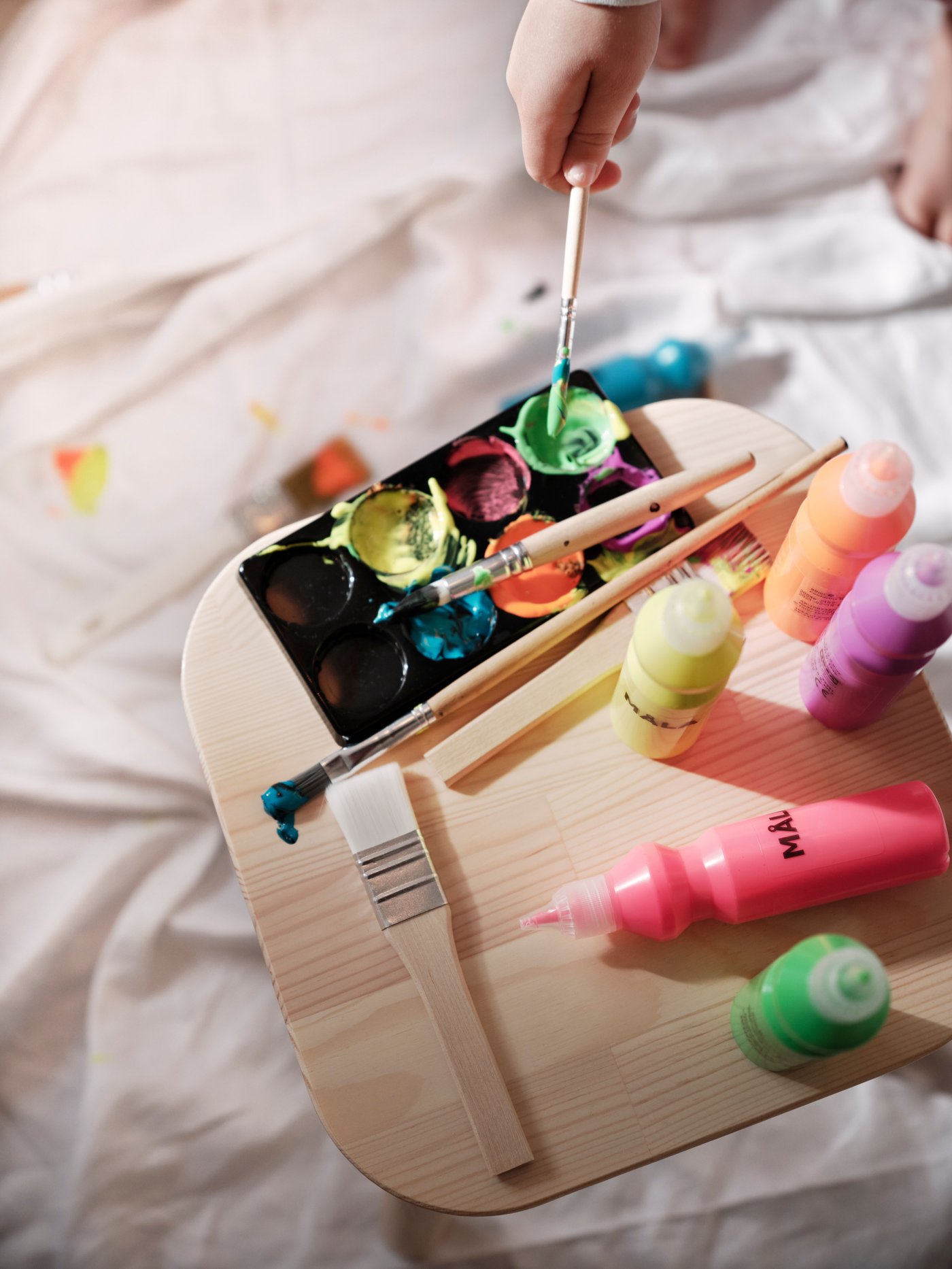 A child’s hand dips a brush into a vibrant palette of paint, surrounded by tubes of mixed colors of IKEA MÅLA fluorescent and glitter paint and brushes on a wooden drawing tray.