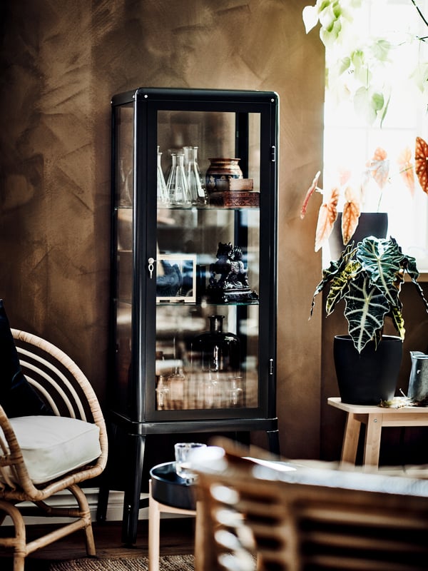 Gemütlicher Boho-chic kombiniert bequeme Korbsessel mit erdigen Tönen und Pflanzen. Eine Glasvitrine im Hintergrund ist mit verschiedenen Dekorationen und Sammlerstücken gefüllt.