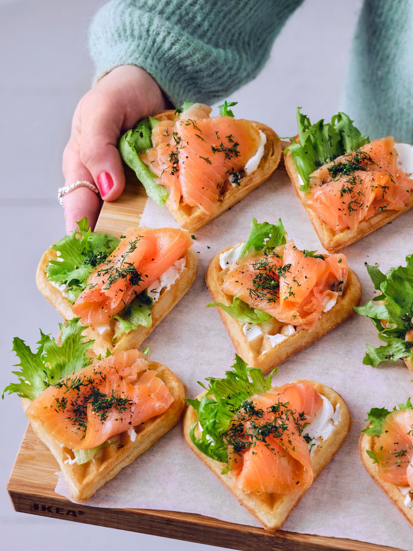 Une personne vêtue d'un pull bleu tient une planche en bois sur laquelle se trouvent douze gaufres avec du saumon, du fromage frais et de la laitue.