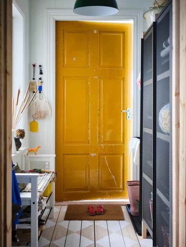 Dans un vestibule, banc avec rangement pour chaussures HEMNES blanc et armoires IVAR grises avec portes en grillage. Porte extérieure jaune vif entrebâillée.