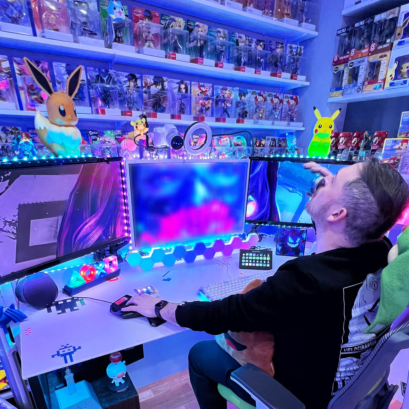 A man is sitting at a table, drinking soda from a can, and playing videogames on three big computer screens. The walls around him are covered with open shelves full of videogames and toys.