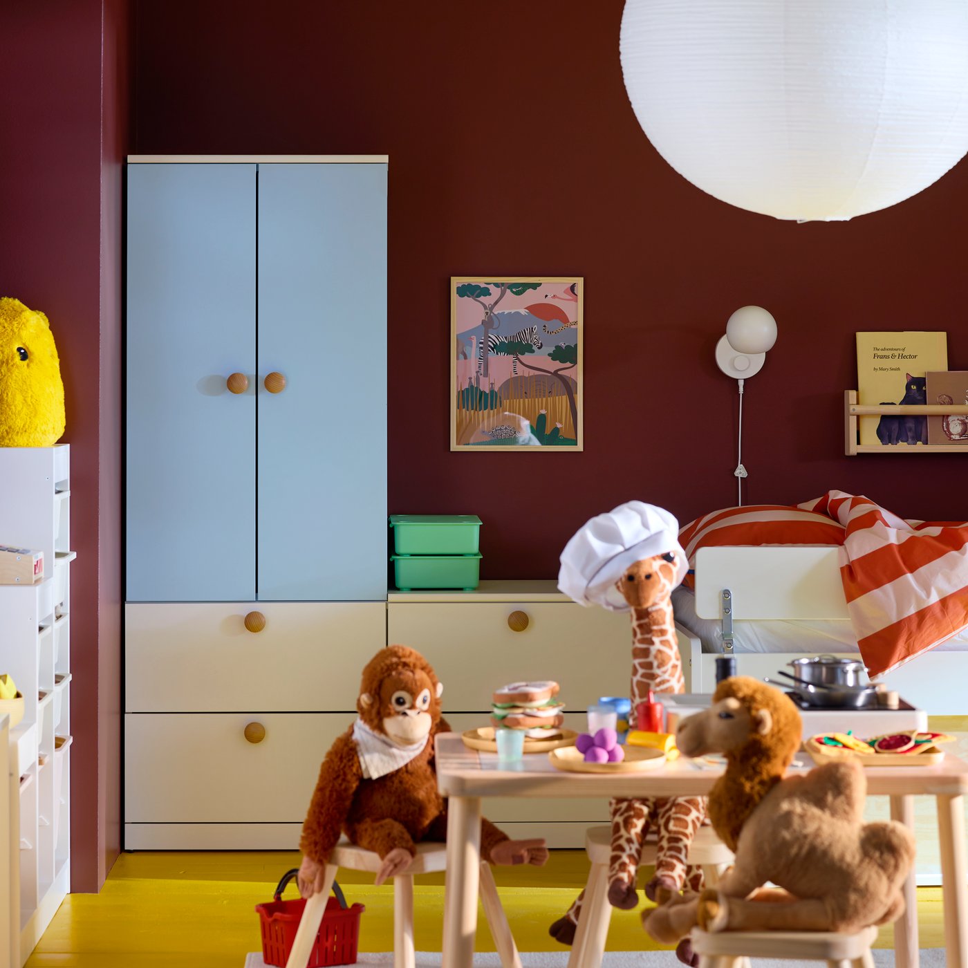 A light wooden FLISAT table in a children’s room with soft toys cooking food, a blue LASTARE wardrobe and a brown-red wall.