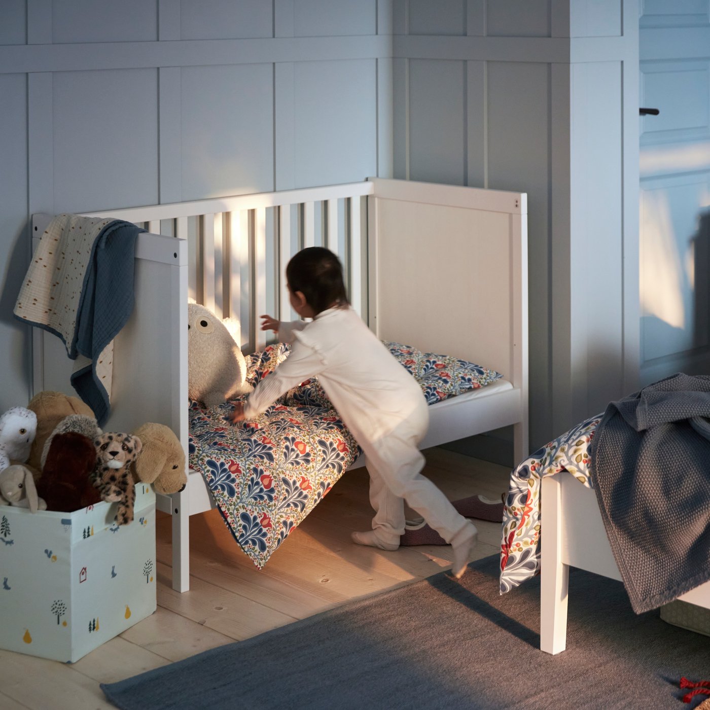 A child in white pyjamas is reaching for a soft toy inside a white SUNDVIK wooden cot with an open long side.