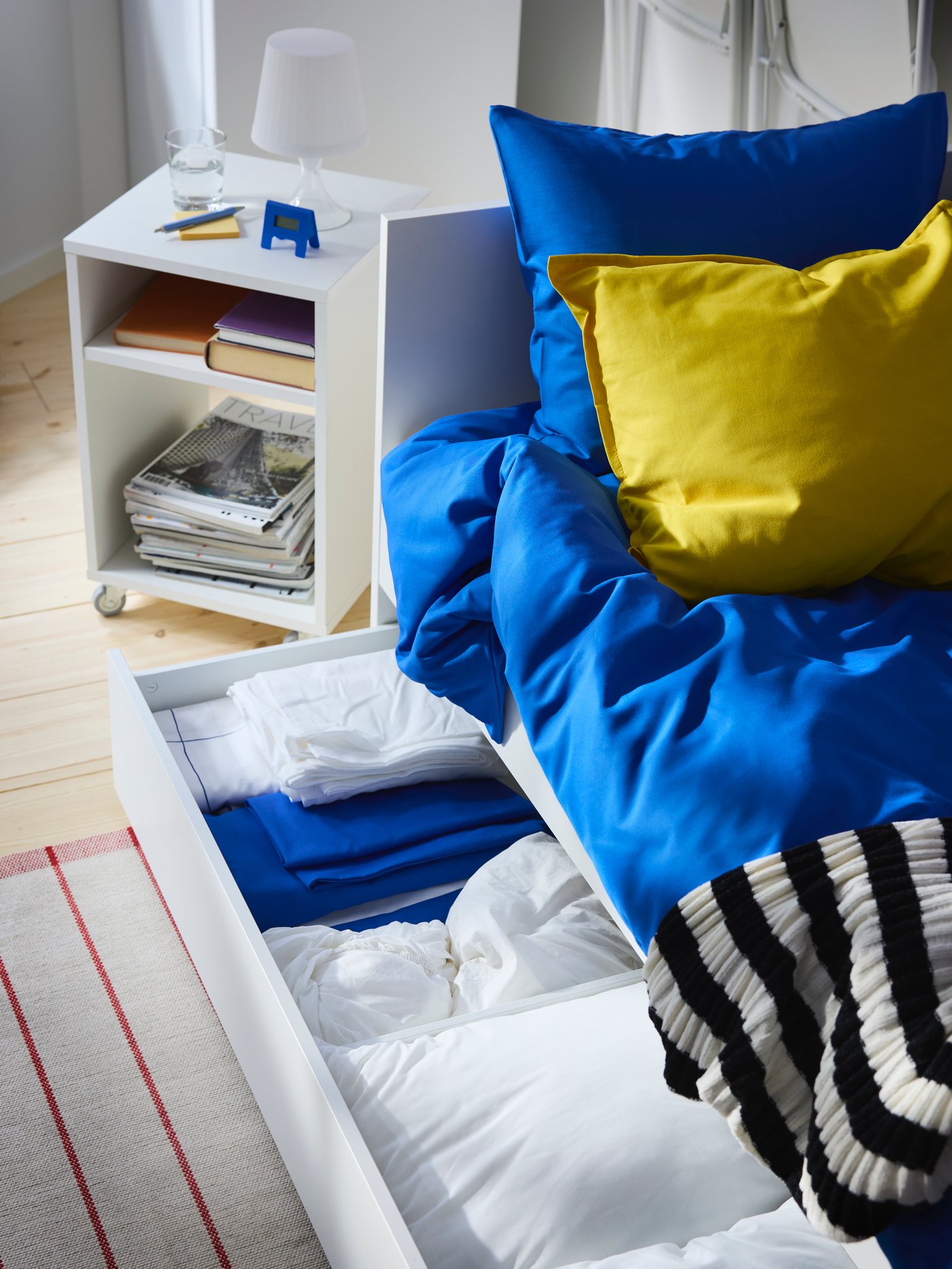 Die Schublade unter dem VIHALS-Tagesbett lässt sich herausziehen und bietet Platz für zusätzliche Bettwäsche. Das Bett ist in leuchtendem Blau und Gelb bezogen.