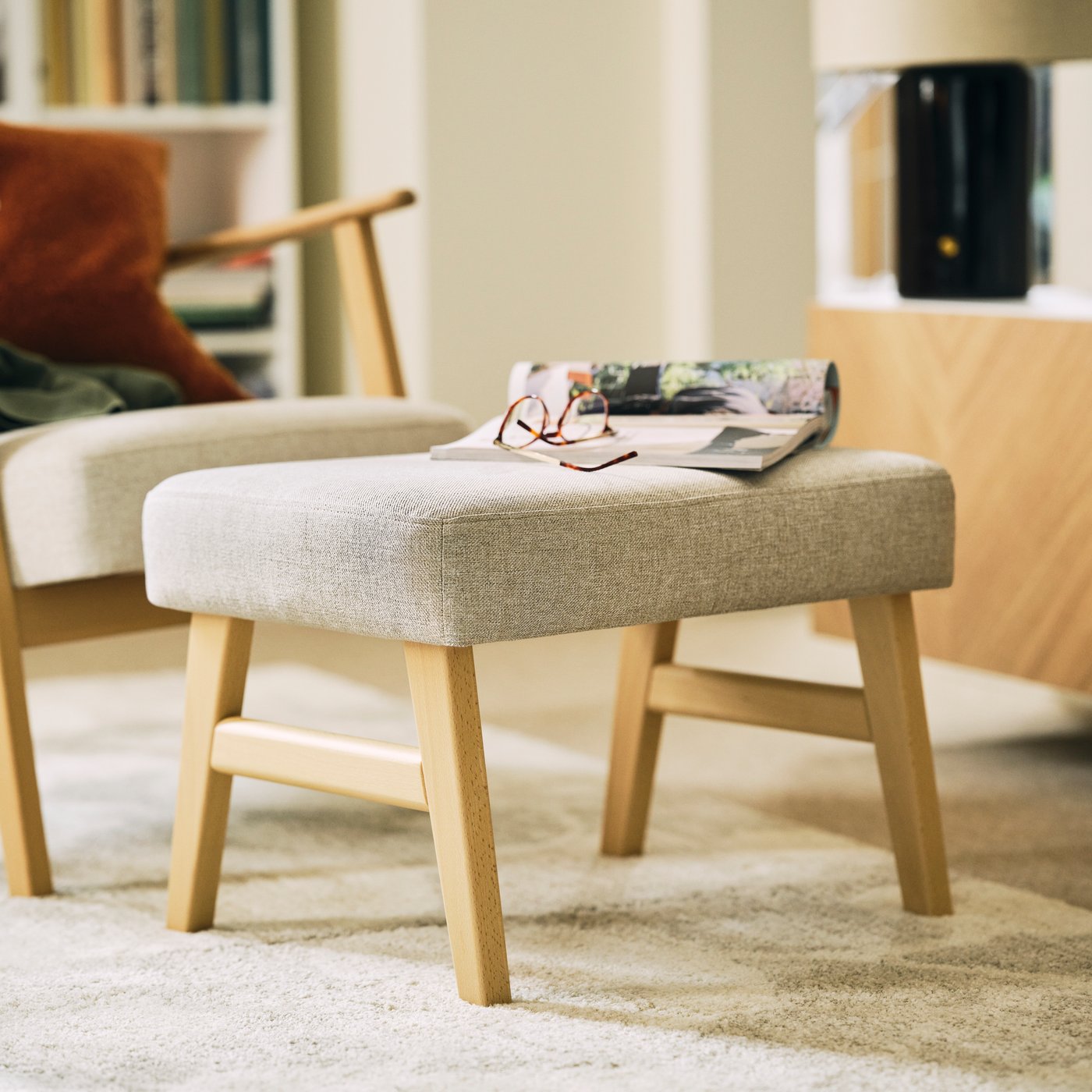 An open magazine and a pair of glasses resting on a light-beige EKENÄSET footstool next to an EKENÄSET armchair.