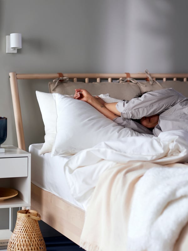Seseorang berpakaian pijama sedang baring di atas katil BJÖRKSNÄS dengan linen katil berwarna putih dan alas ODDRUN, di sebelah meja sisi katil berwarna putih.