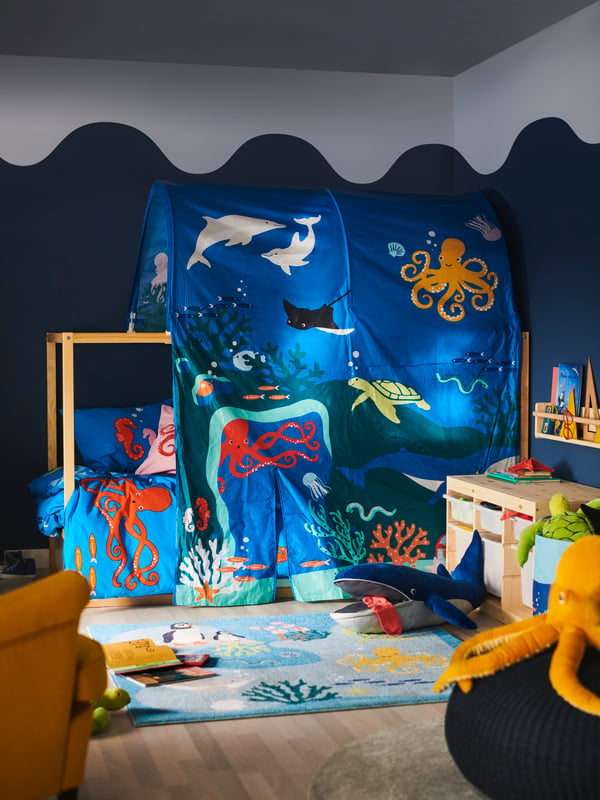 A bed is covered in a KURA ocean animals bed tent; a BLÅVINGAD blue whale soft toy is on a BLÅVINGAD rug in front of it.