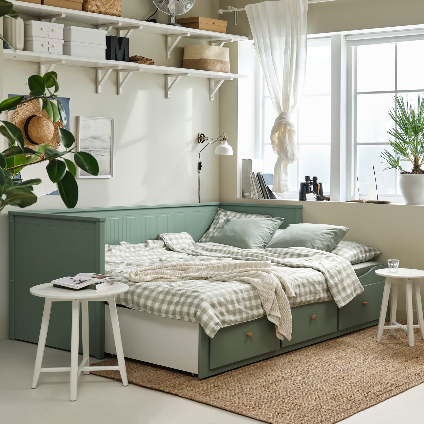 Cozy bedroom with a green daybed, soft bedding, plants, and sunlight through large windows.