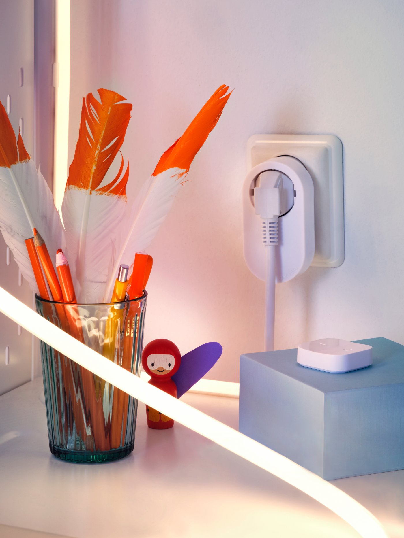 The corner of a small desk with a TRÅDFRI control outlet kit, a LED lighting strip and a glass holding pencils and feathers.