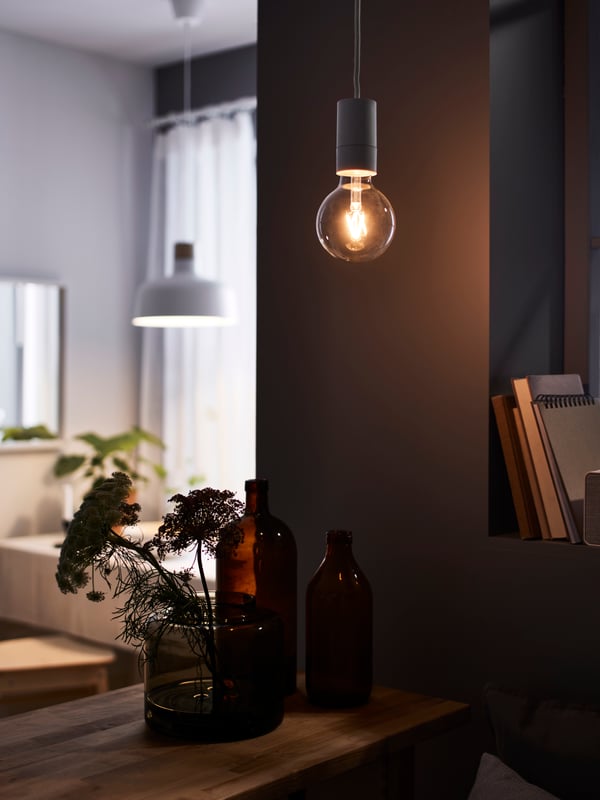 LUNNOM and SUNNERBY naked bulb with cord hanging above a FORHOJA side cabinet with a KONSTFULL vase of flowers on top. A BUNKEFLO pendant lamp is seen in the background above a sofa  in the living room. 