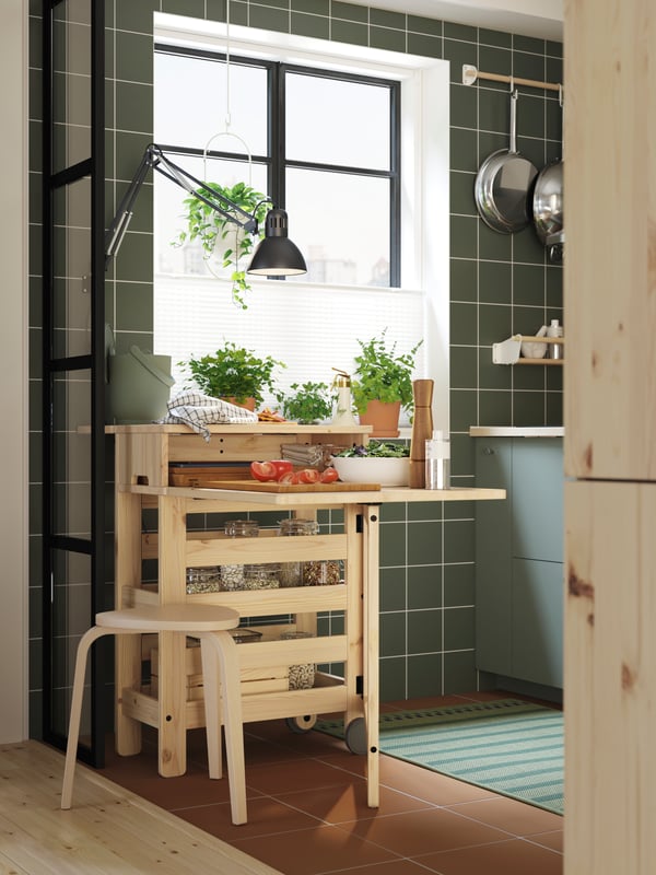 A RESARÖ mobile drop-leaf table with storage is placed below a window. It’s folded out and has a wooden stool next to it.