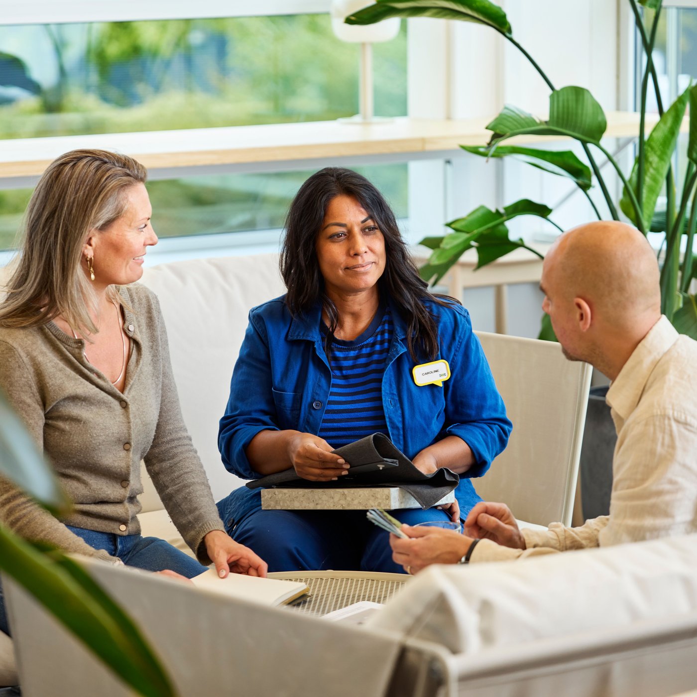 An IKEA interior designer with fabric samples in her hand sits on a sofa with two customers and advises them.