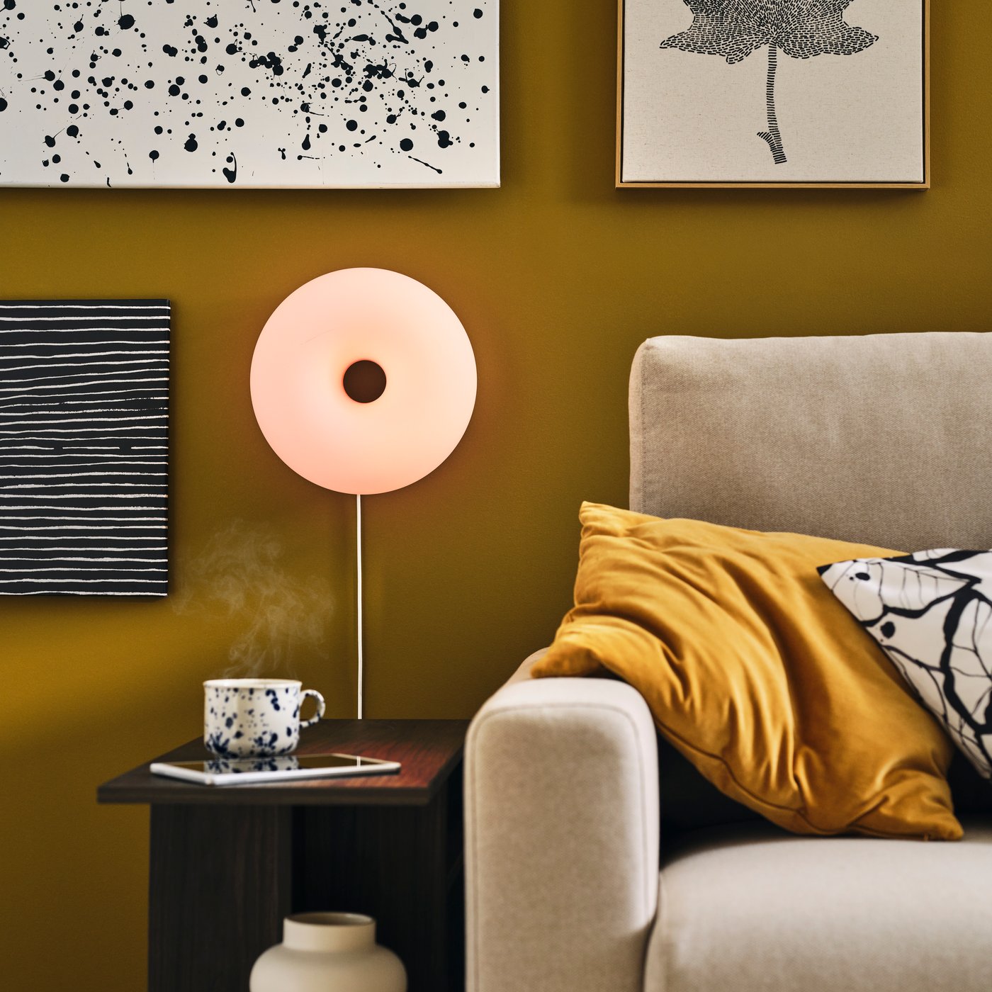 A lit VARMBLIXT table-wall lamp flanked by art and hung over a HOLMERUD side table next to a light-beige MALMKÖPING sofa.