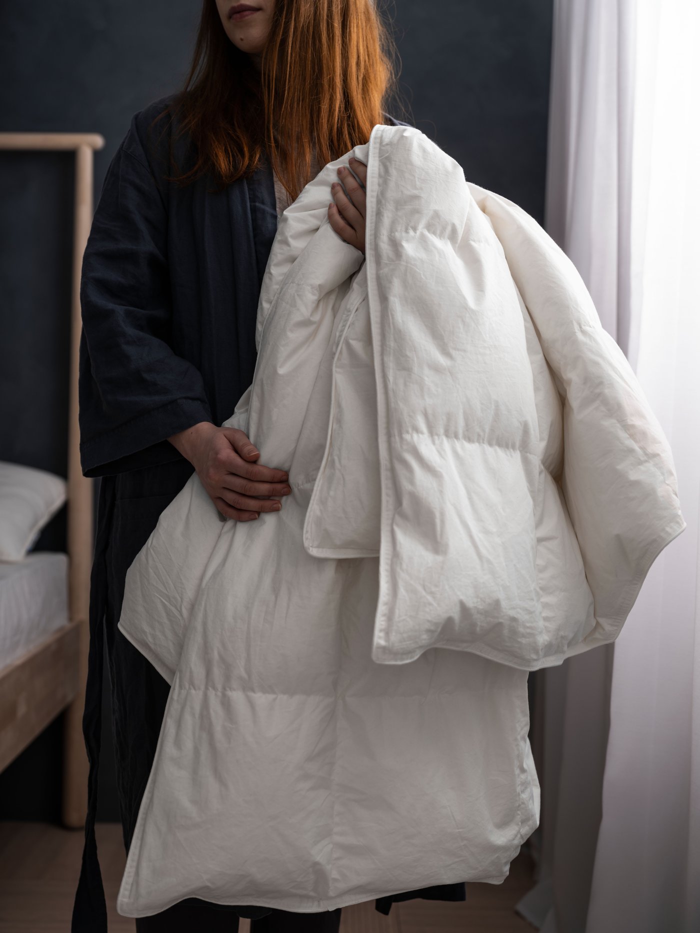 A person holds a large, white duvet in a bedroom with dark walls and a light curtain.