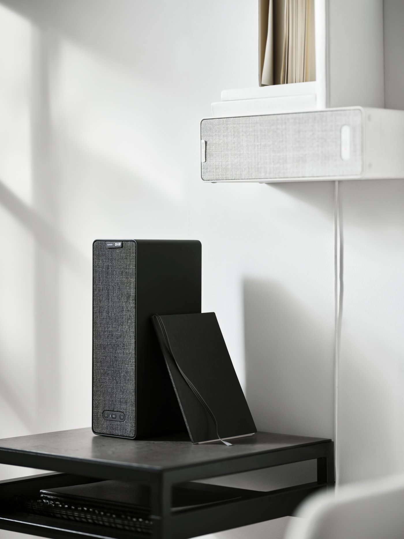Two SYMFONISK WiFi bookshelf speakers, the white one is on the wall with books on it, the black one is on a black side table.